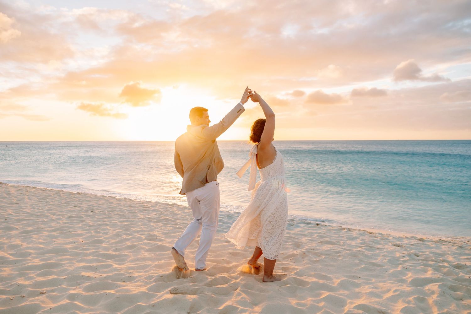 Couple dancing on a white sandy beach at sunset, holding hands raised, with turquoise ocean waves in the background.