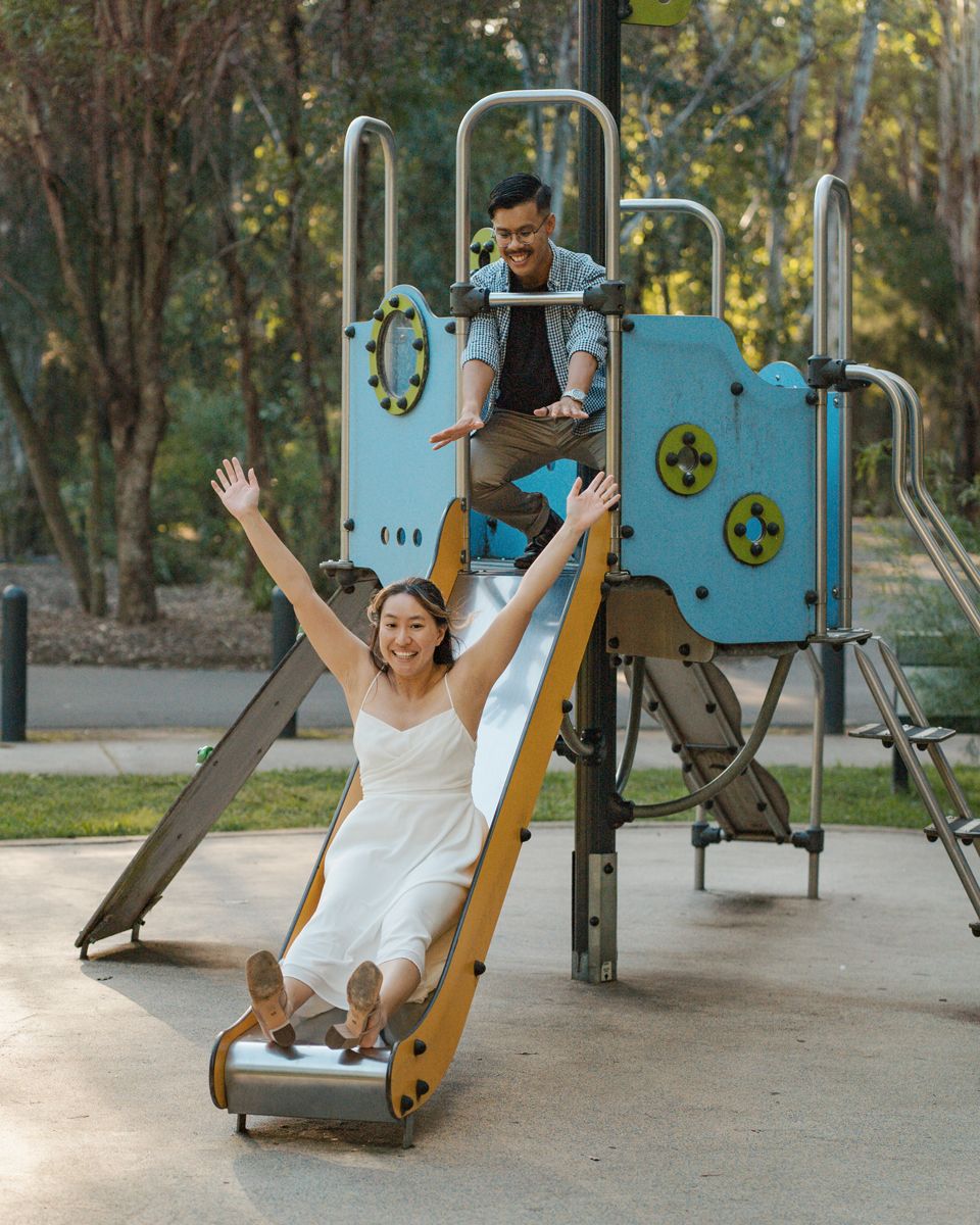 Couple in wedding attire playing on a playground slide in a park, bride raising arms joyfully during a photoshoot in western sydney parkland
