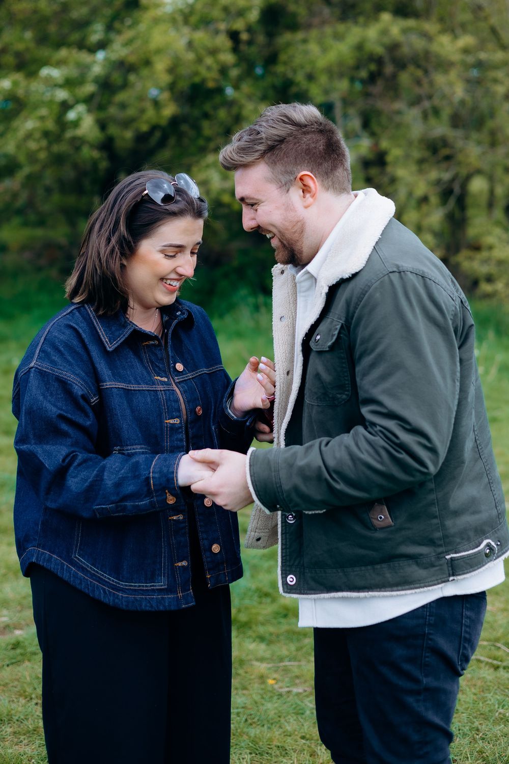 A couple laughing and holding hands outdoors, woman in denim jacket, man in green coat, green trees behind them.