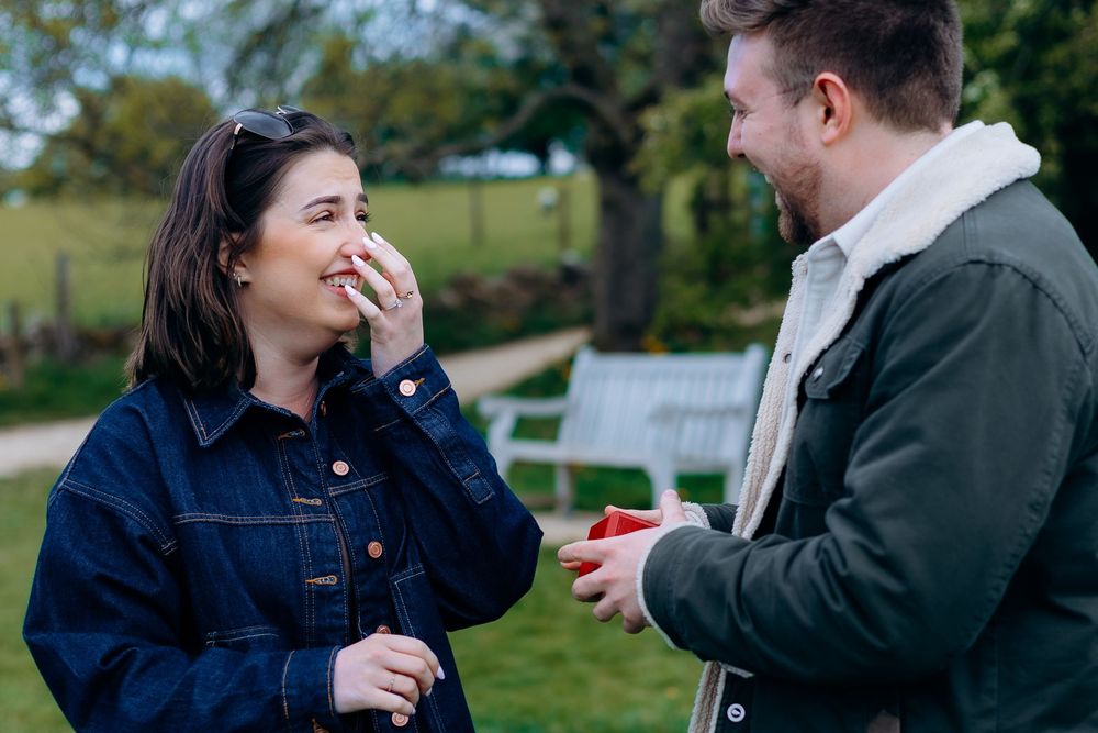Man proposing to surprised woman outdoors, holding ring box while she covers her mouth in joy.