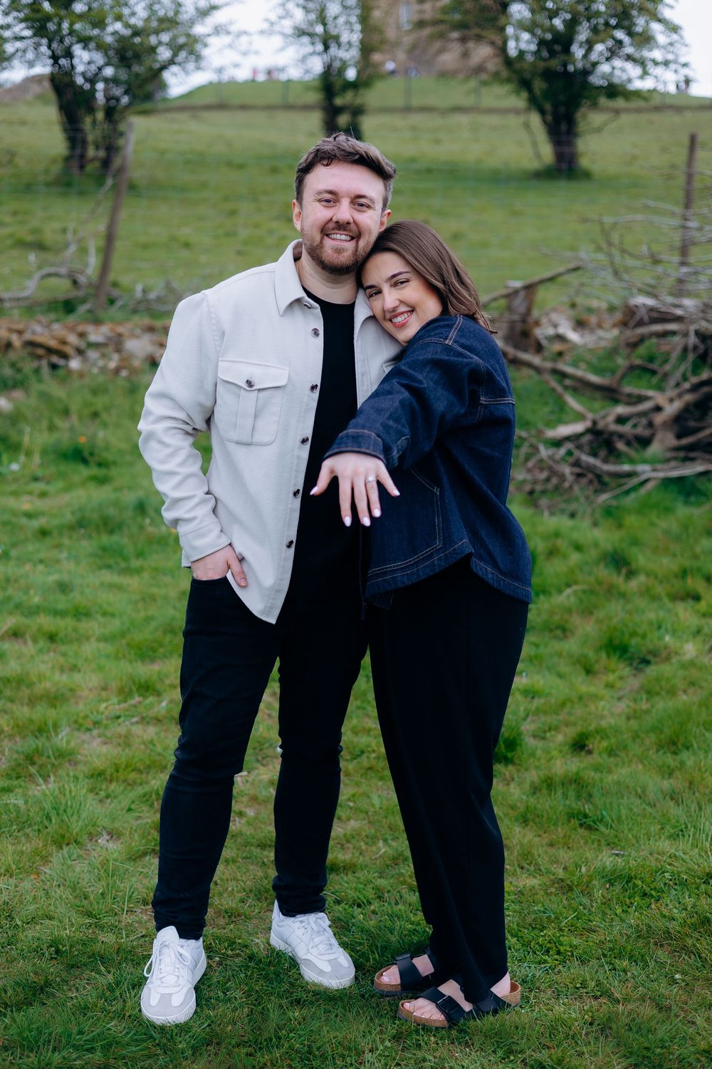 A smiling couple poses together on a green grassy field, the woman leaning on the man's shoulder outdoors.