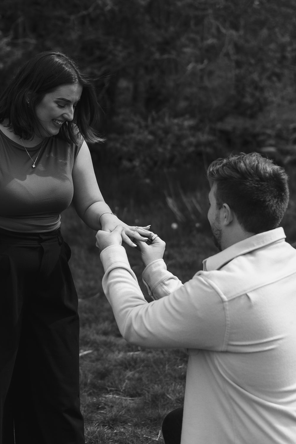 Black and white photo of a man on one knee proposing to a smiling woman outdoors.