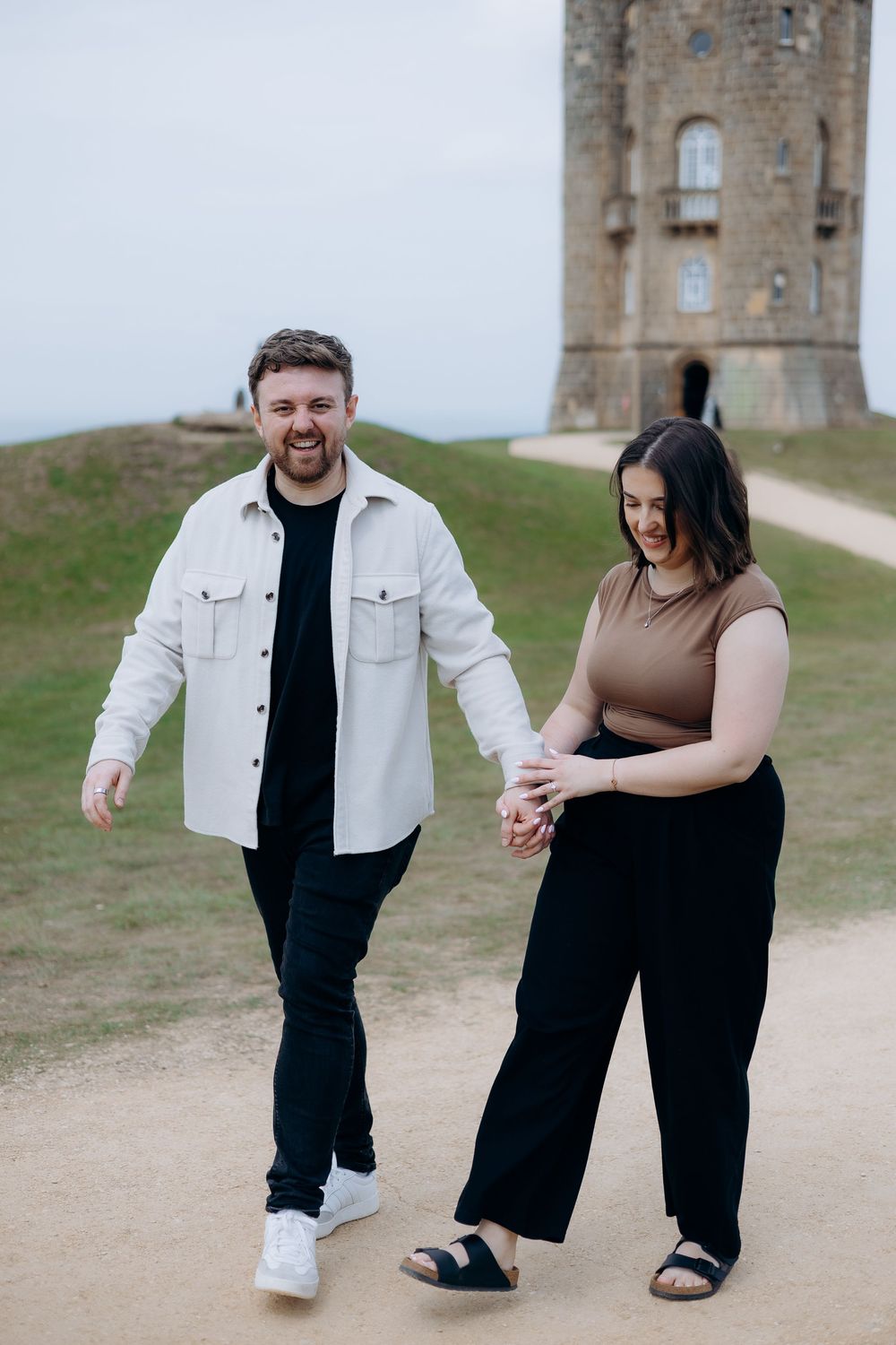 Couple holding hands and walking on a path near a historic stone tower on a grassy hill.