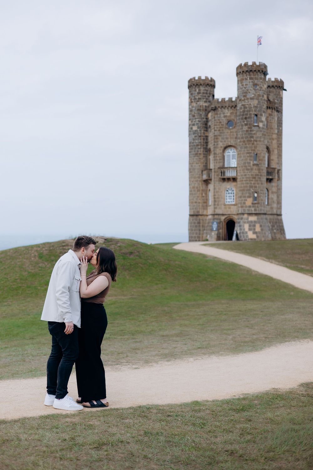 Couple sharing a romantic kiss on a winding path with Broadway Tower castle in the background on a cloudy day.
