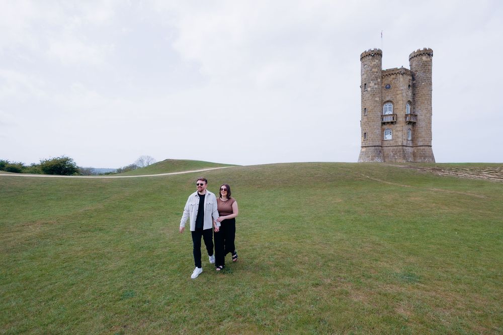 Couple standing on green hill before Broadway Tower, a historic folly in the Cotswolds, England.