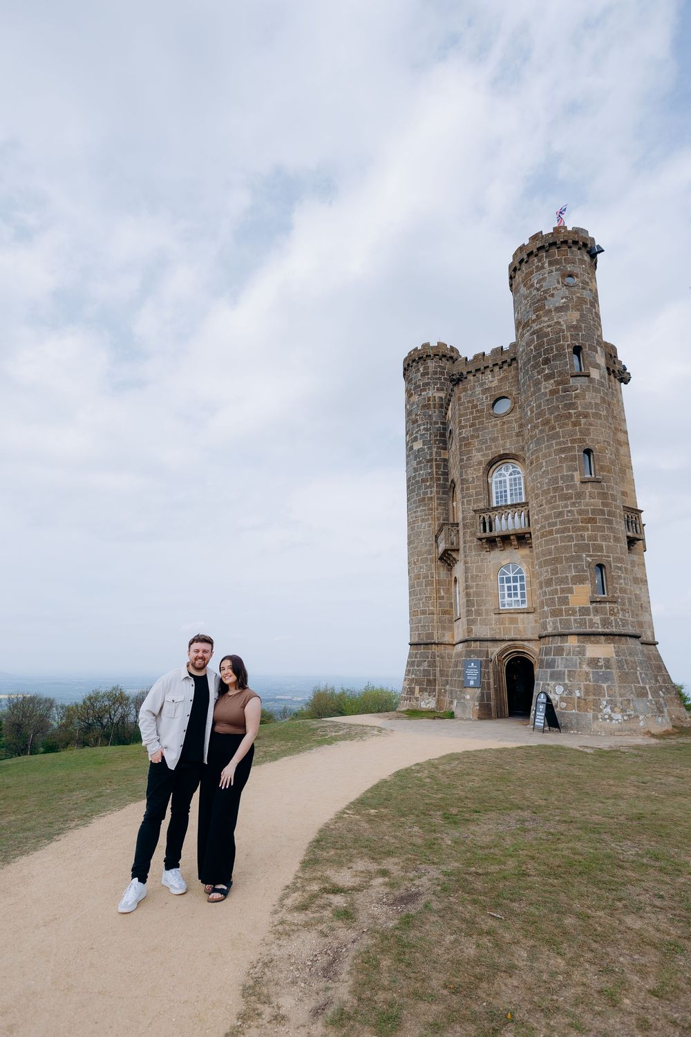 Couple posing together on a path beside Broadway Tower, a historic folly castle in the Cotswolds, England.
