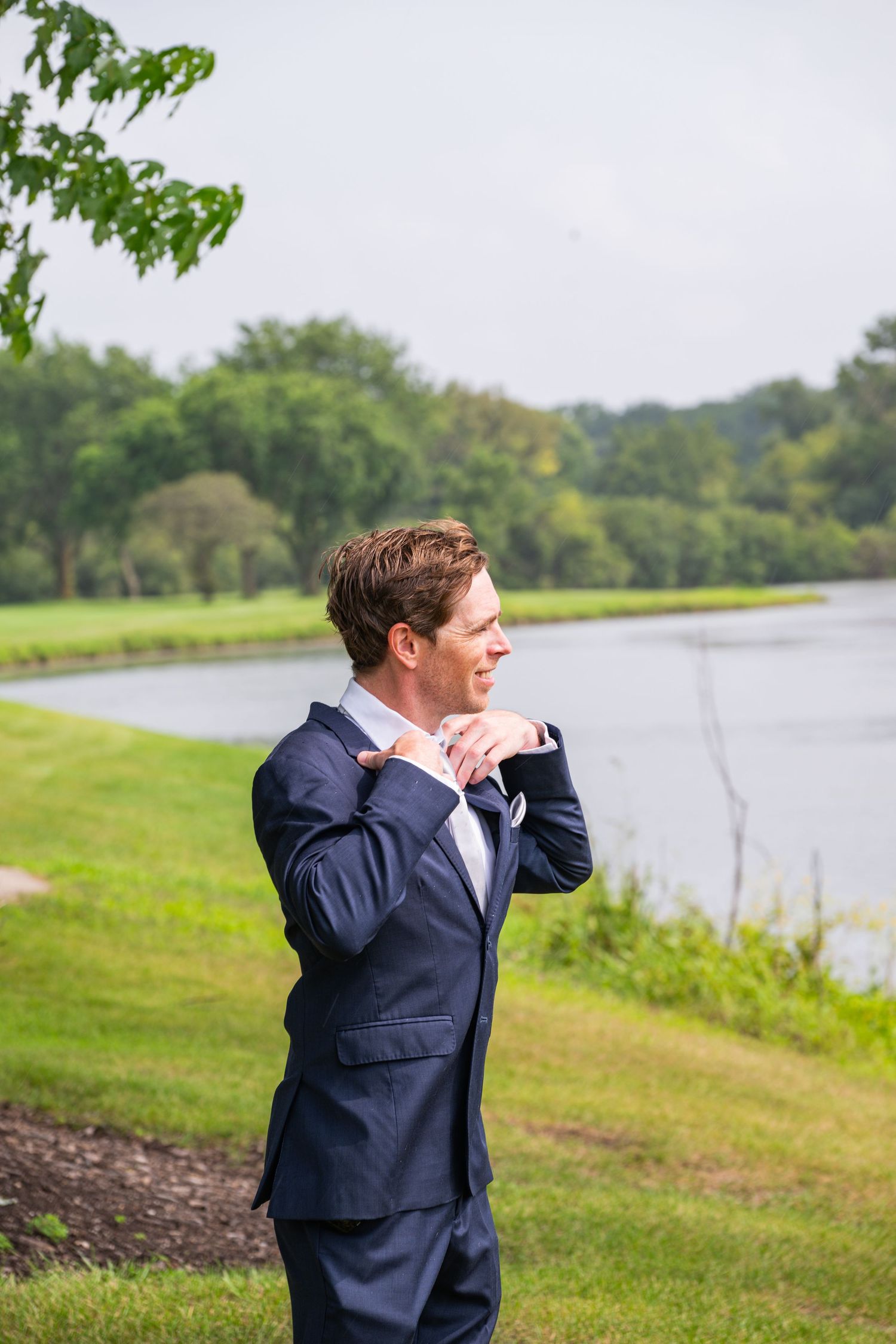 A groom in a navy blue suit adjusts his tie outdoors near a serene pond surrounded by lush green trees.