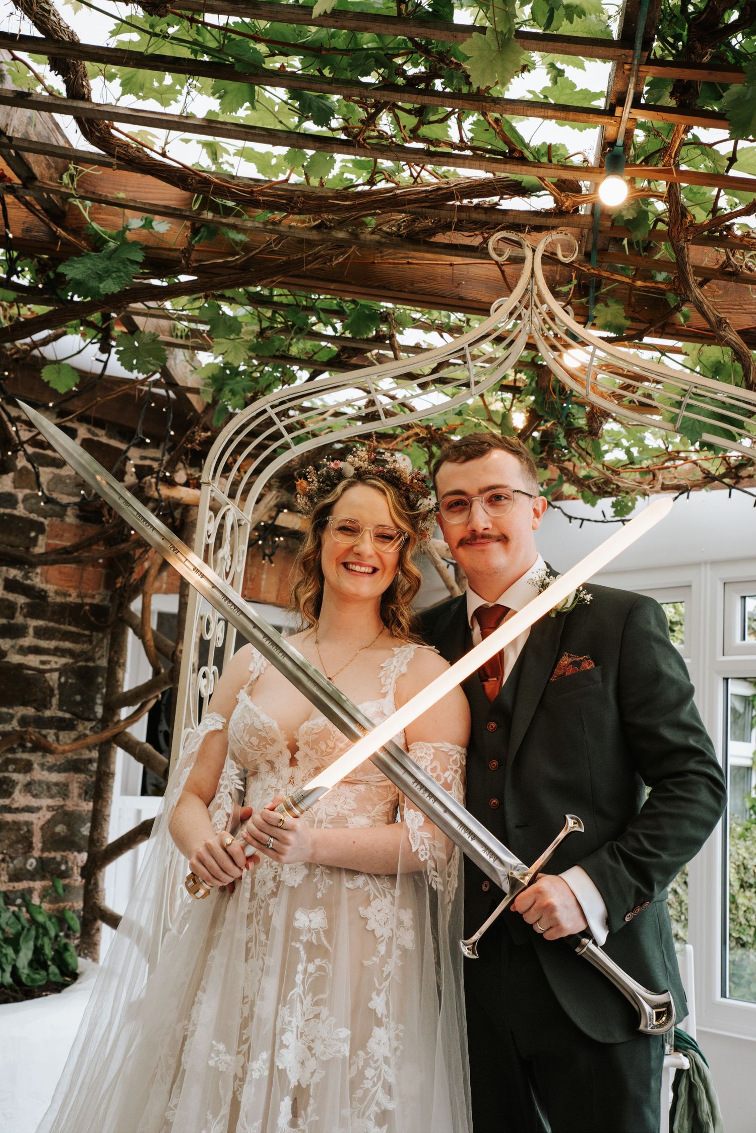 Bride and groom holding crossed swords at their fantasy-themed wedding under a vine-covered pergola.