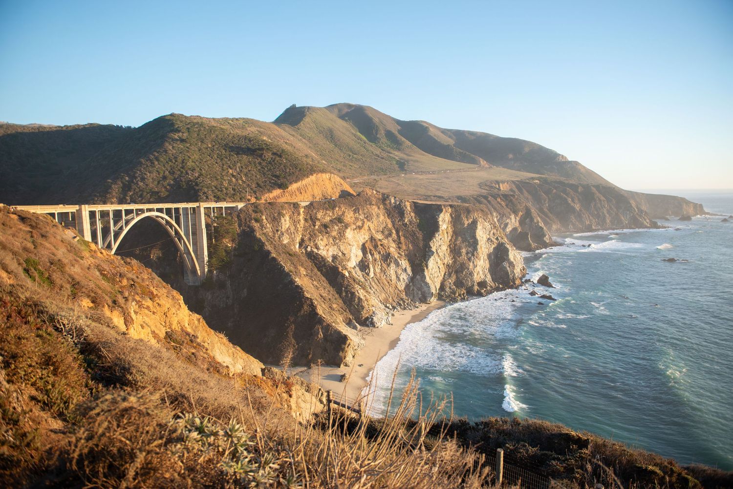 Bixby Creek Bridge arches over rugged Big Sur coastline with Pacific Ocean waves and golden cliffs under clear blue sky.