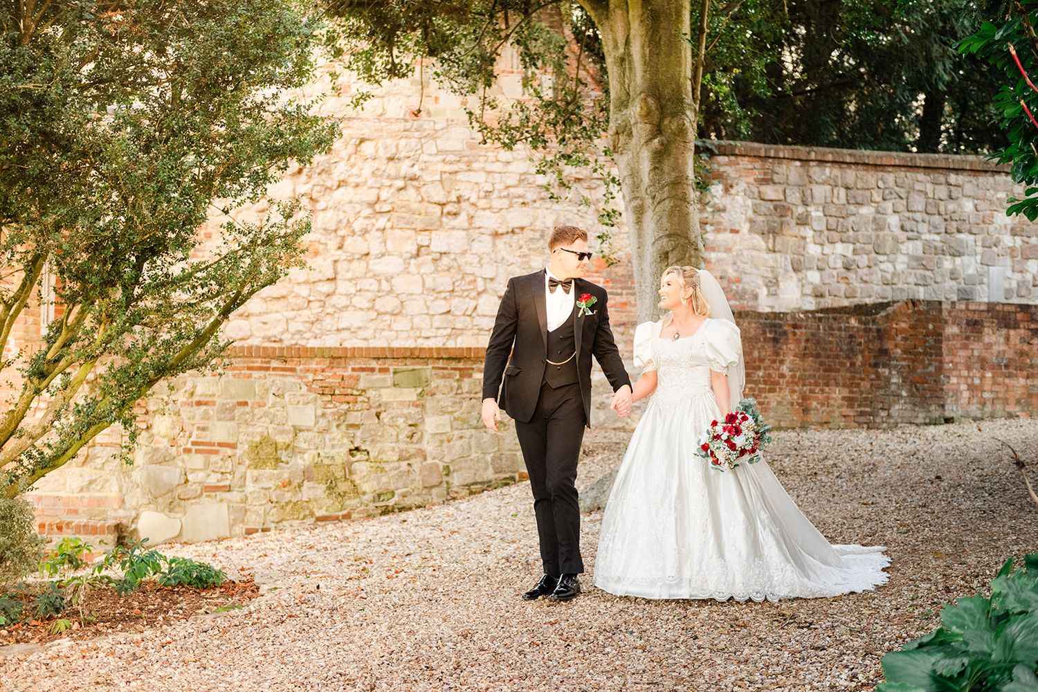 Bride and groom walk hand-in-hand on gravel path beside rustic stone wall surrounded by lush greenery.