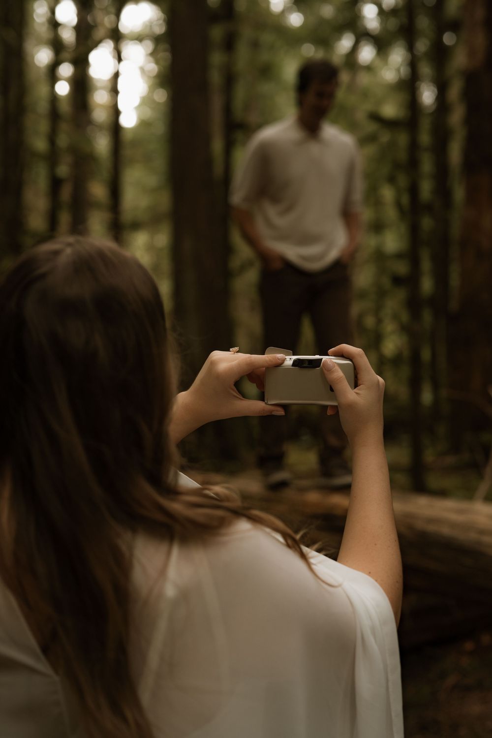 Woman photographs a man standing among tall trees in a moody forest setting.