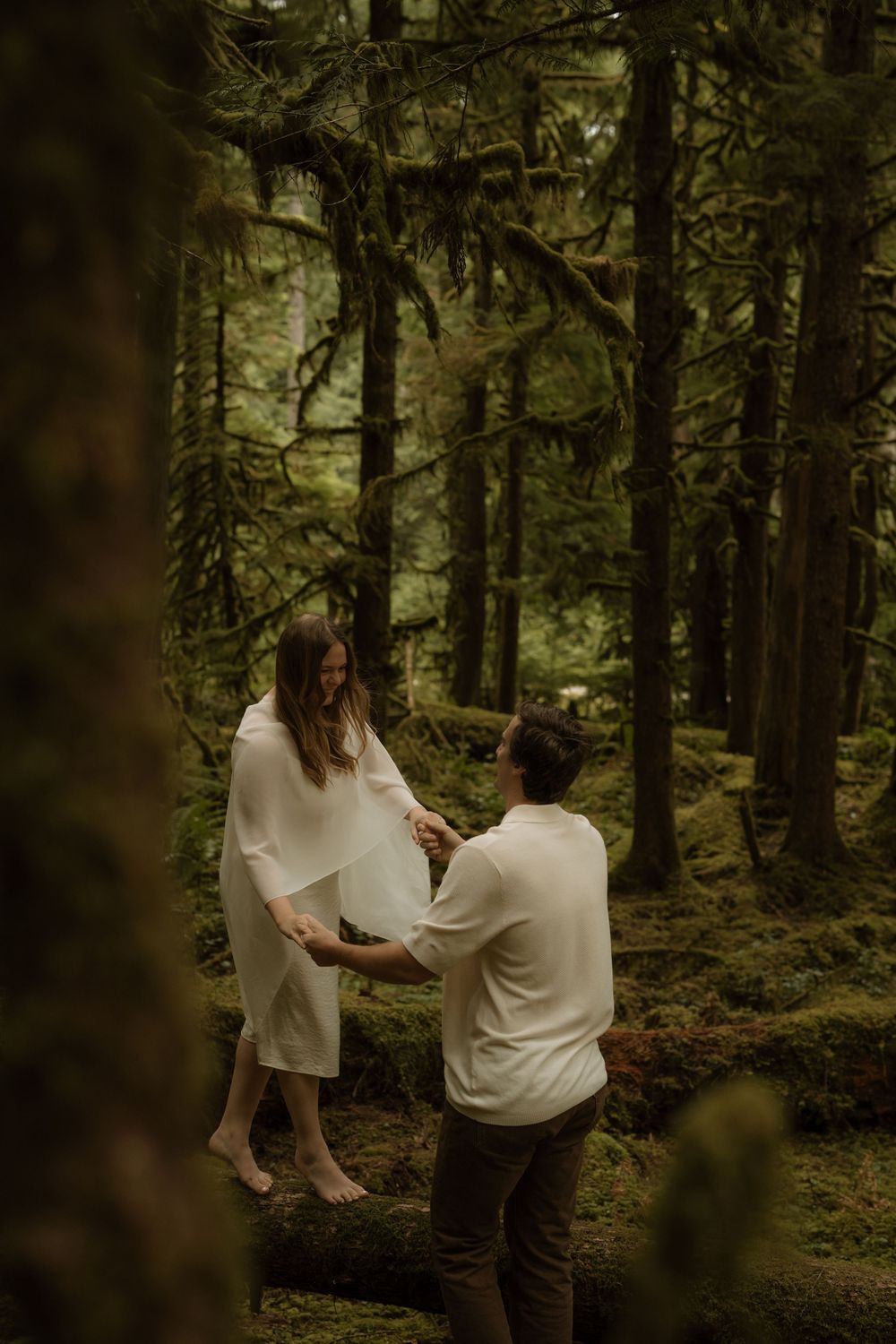 Couple holding hands in a lush mossy forest, woman in white dress standing on log, romantic woodland maternity session.