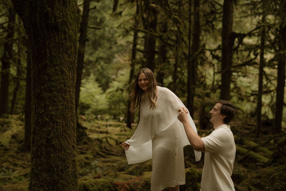 Couple sharing a romantic moment in a lush green forest, woman in flowing white dress laughing as man kneels beside her.