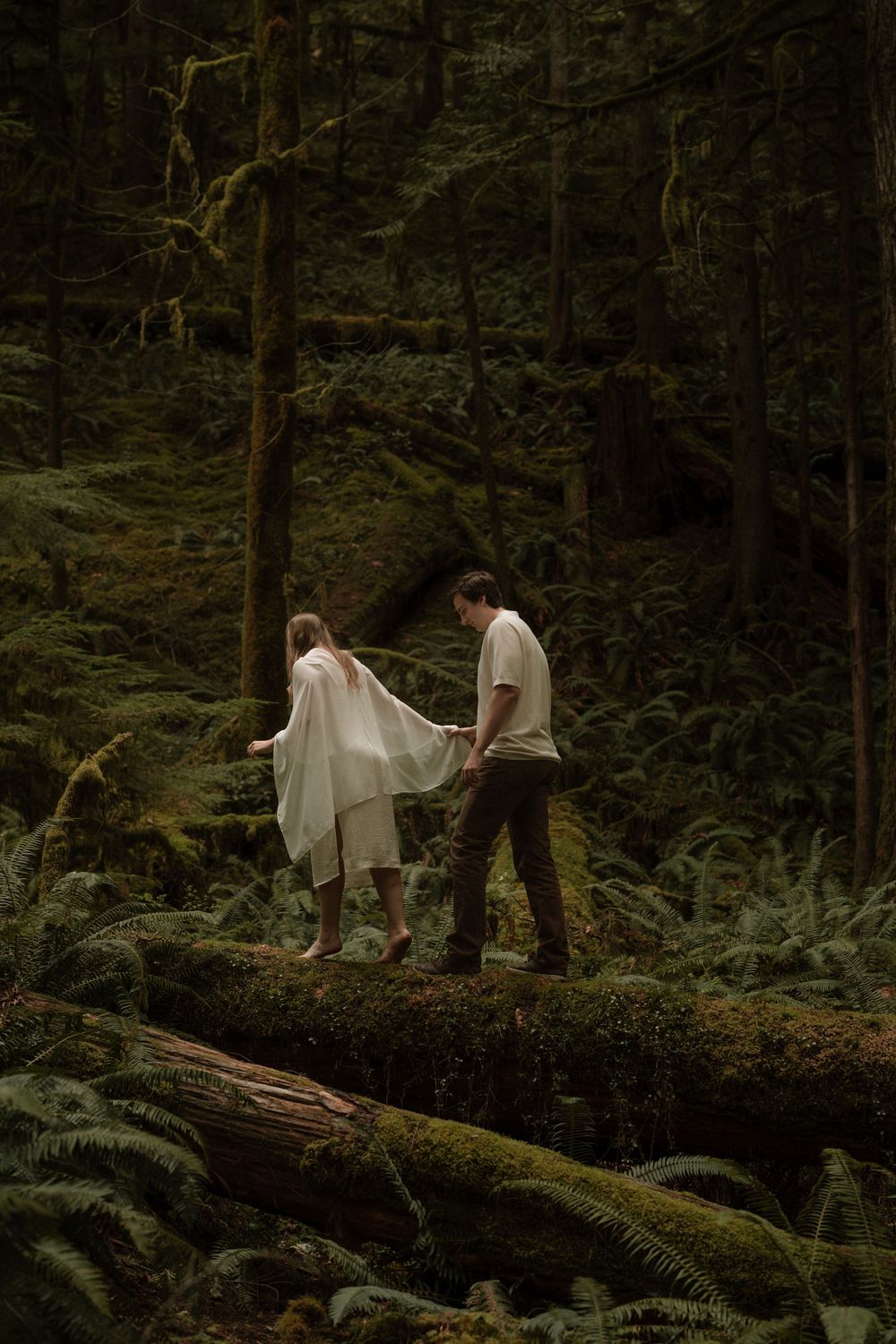 Couple walks on a mossy log in a lush, fern-filled forest.