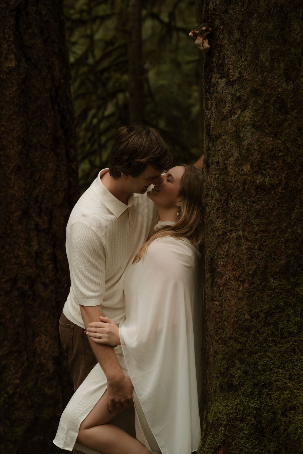 Couple sharing a romantic kiss between moss-covered trees in a lush forest, wearing white outfits.