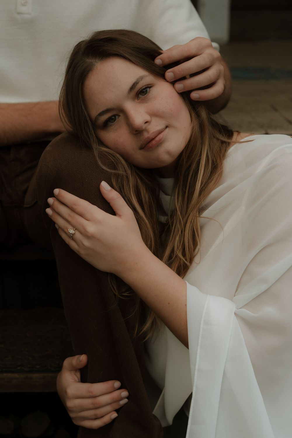 Woman in white blouse leans on man in brown pants, showcasing engagement ring, in an intimate couples portrait.