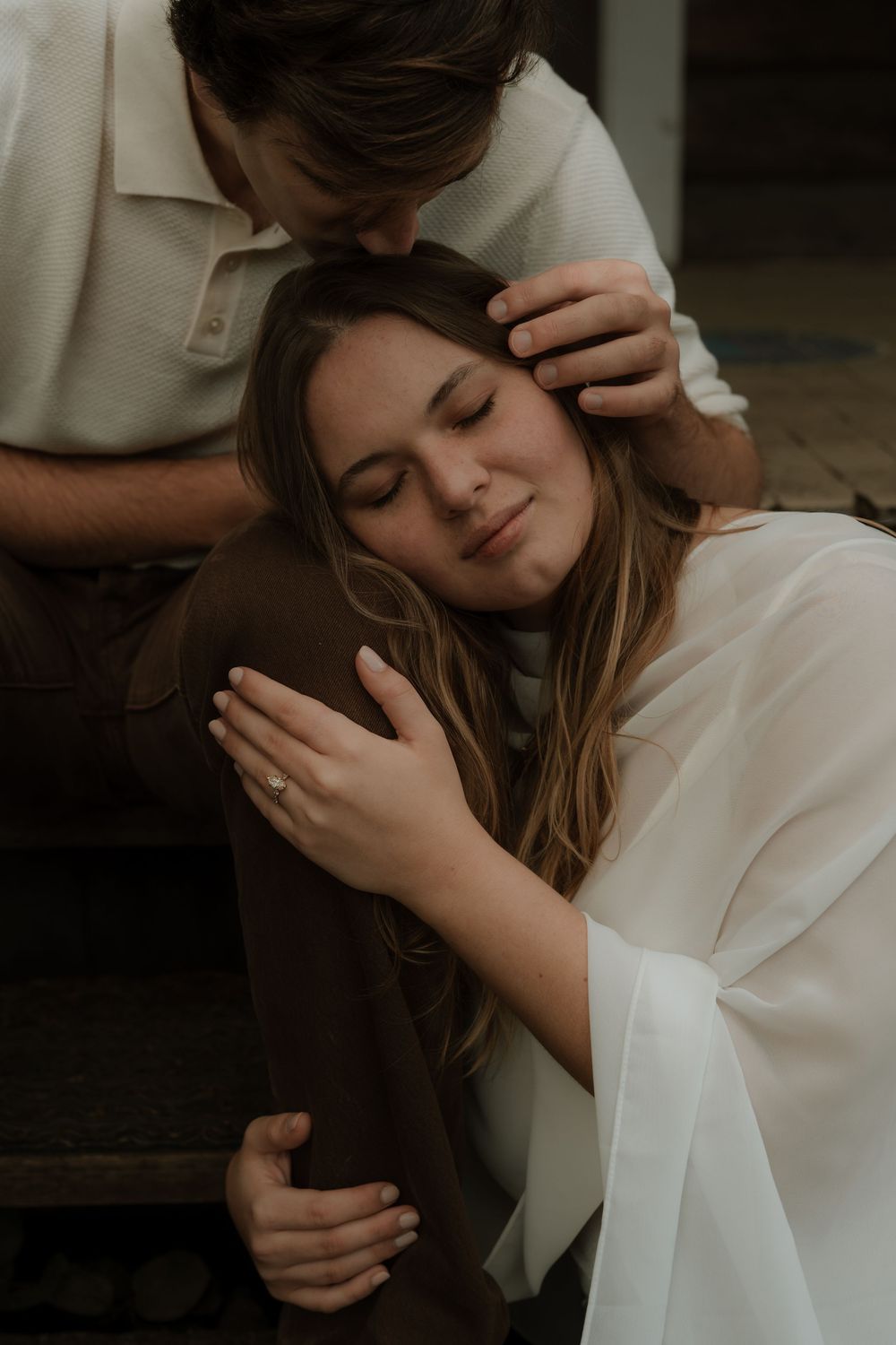 Woman with eyes closed resting her head on her partner's knee while he kisses her forehead tenderly.