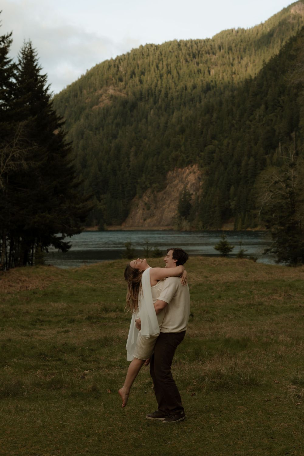 Couple embracing in a meadow with a mountain lake and evergreen forest backdrop during golden hour.