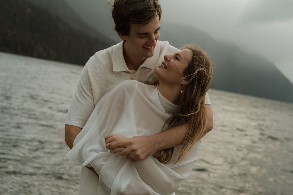 A couple shares a romantic embrace by a misty mountain lake, the man lifting the woman in white.