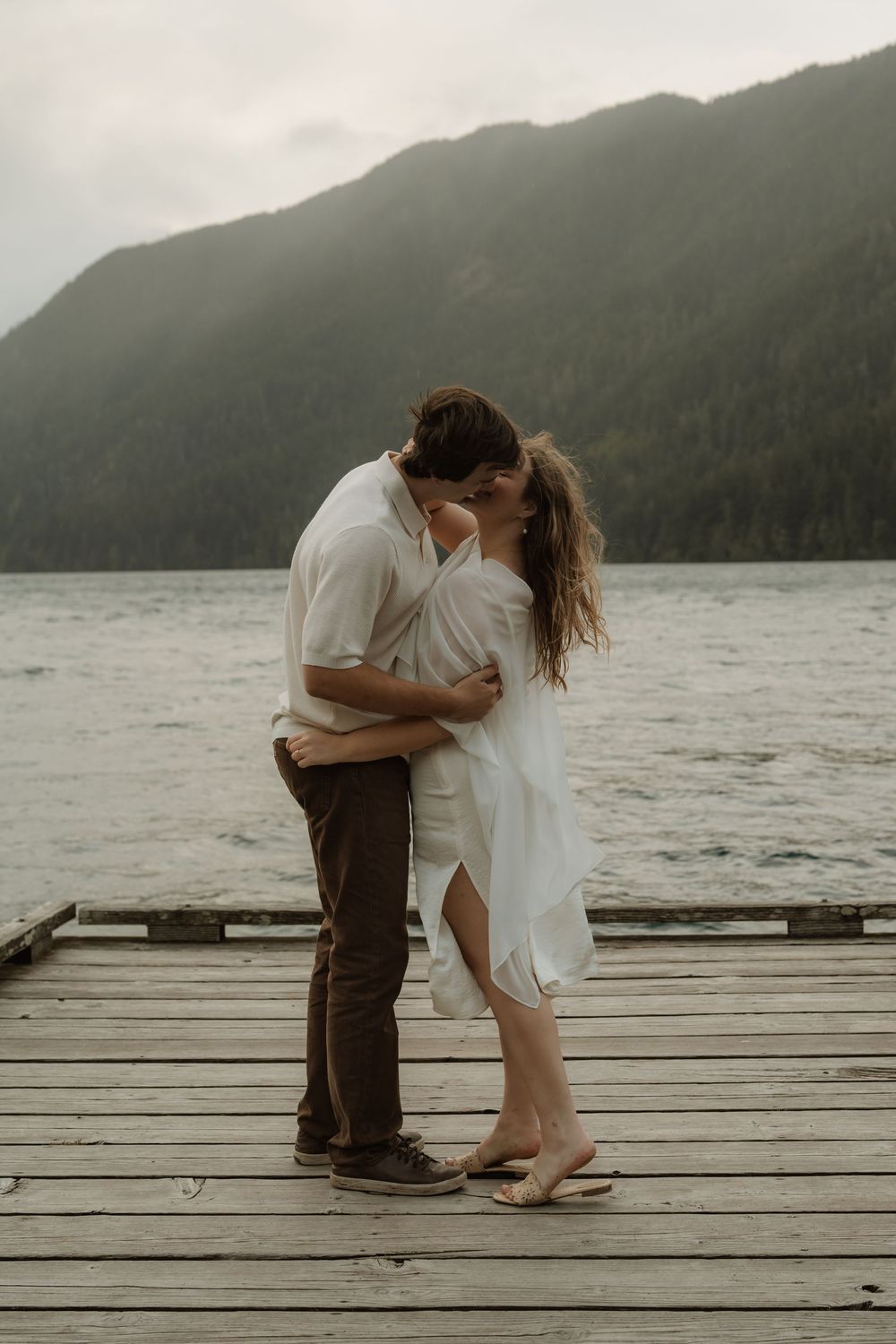 Couple sharing a romantic kiss on a wooden dock with misty mountain lake backdrop.