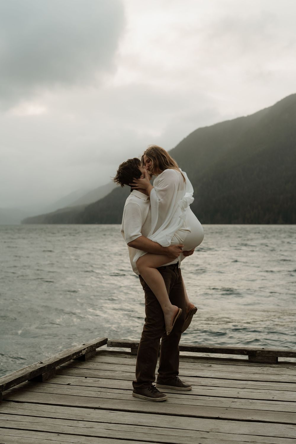 Couple sharing a romantic kiss on a wooden dock overlooking a misty mountain lake on a cloudy day.