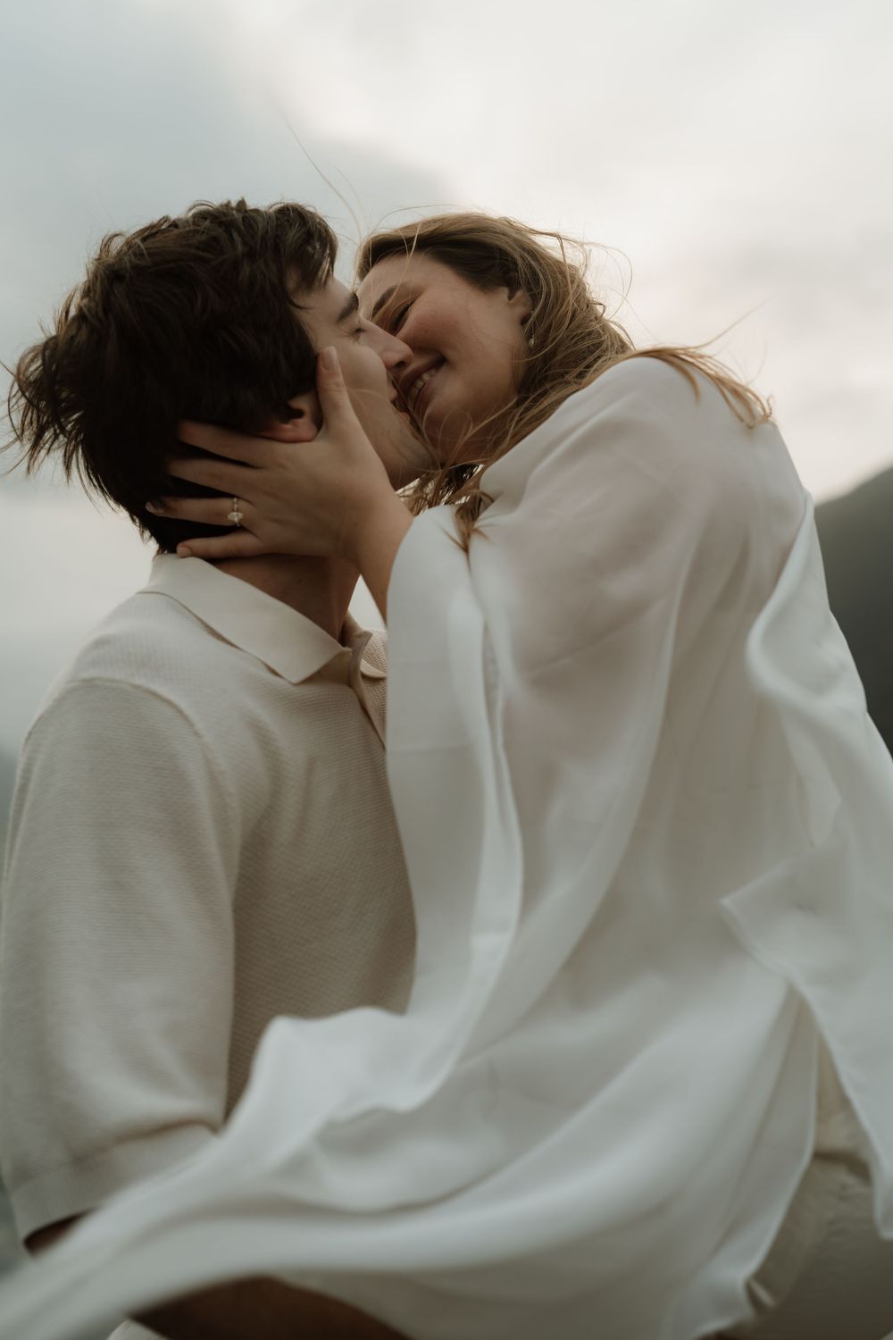 A couple shares a romantic kiss outdoors, dressed in white linen outfits against a cloudy sky backdrop.