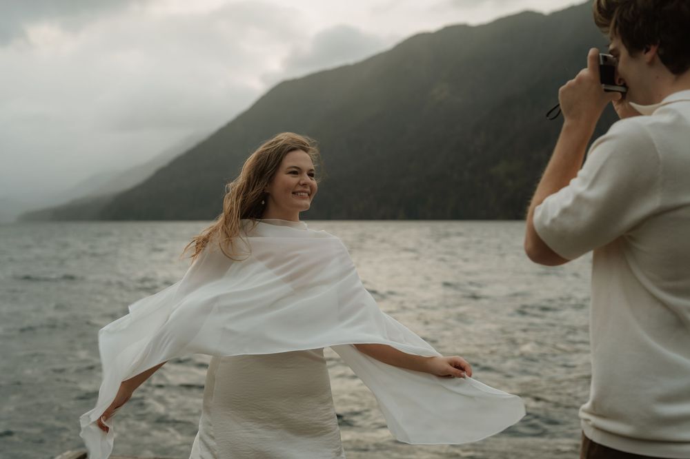 Woman in white wrap laughs as man photographs her by misty mountain lake during outdoor elopement shoot.