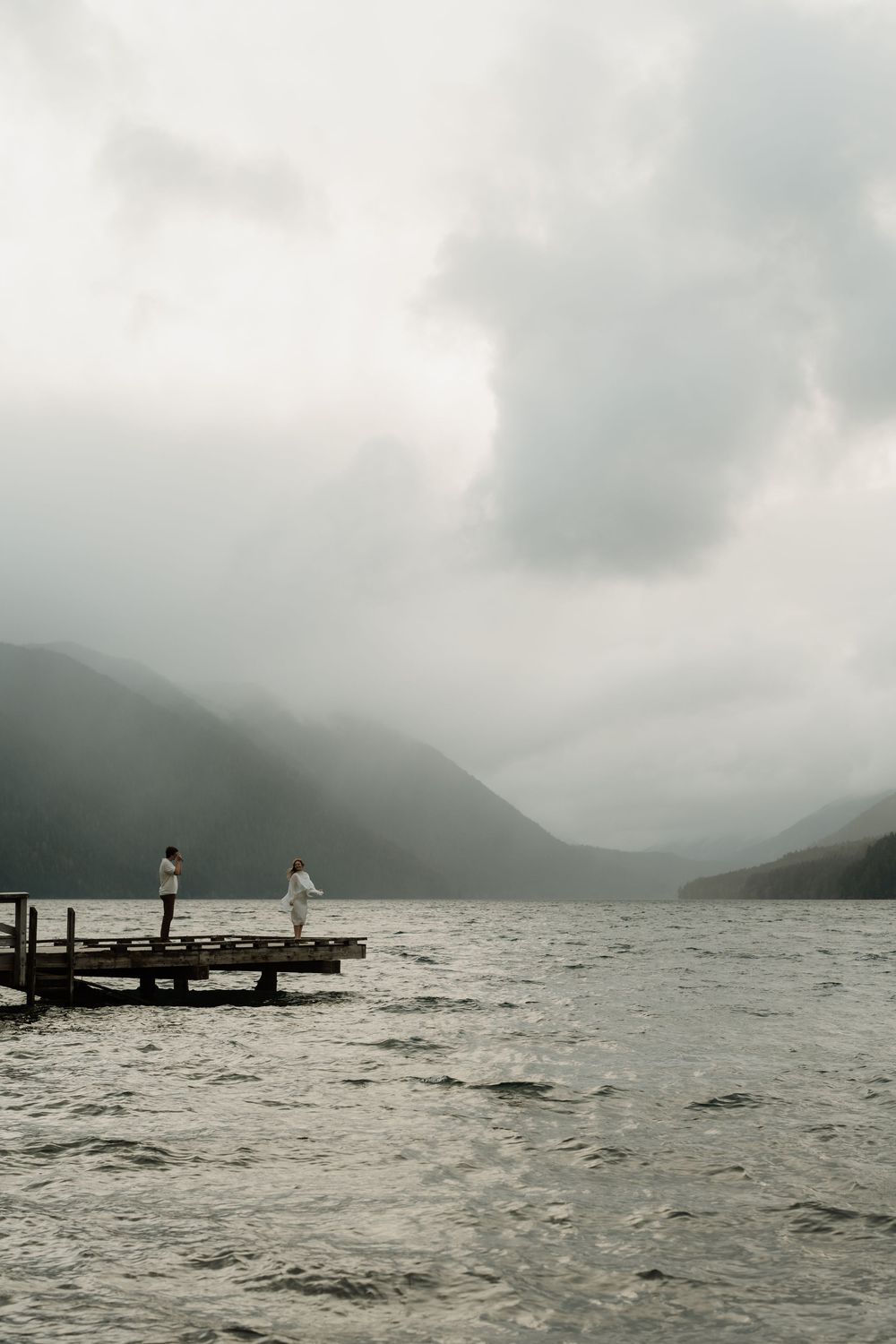 Two people in white stand on a misty lake dock surrounded by mountains under dramatic cloudy skies.