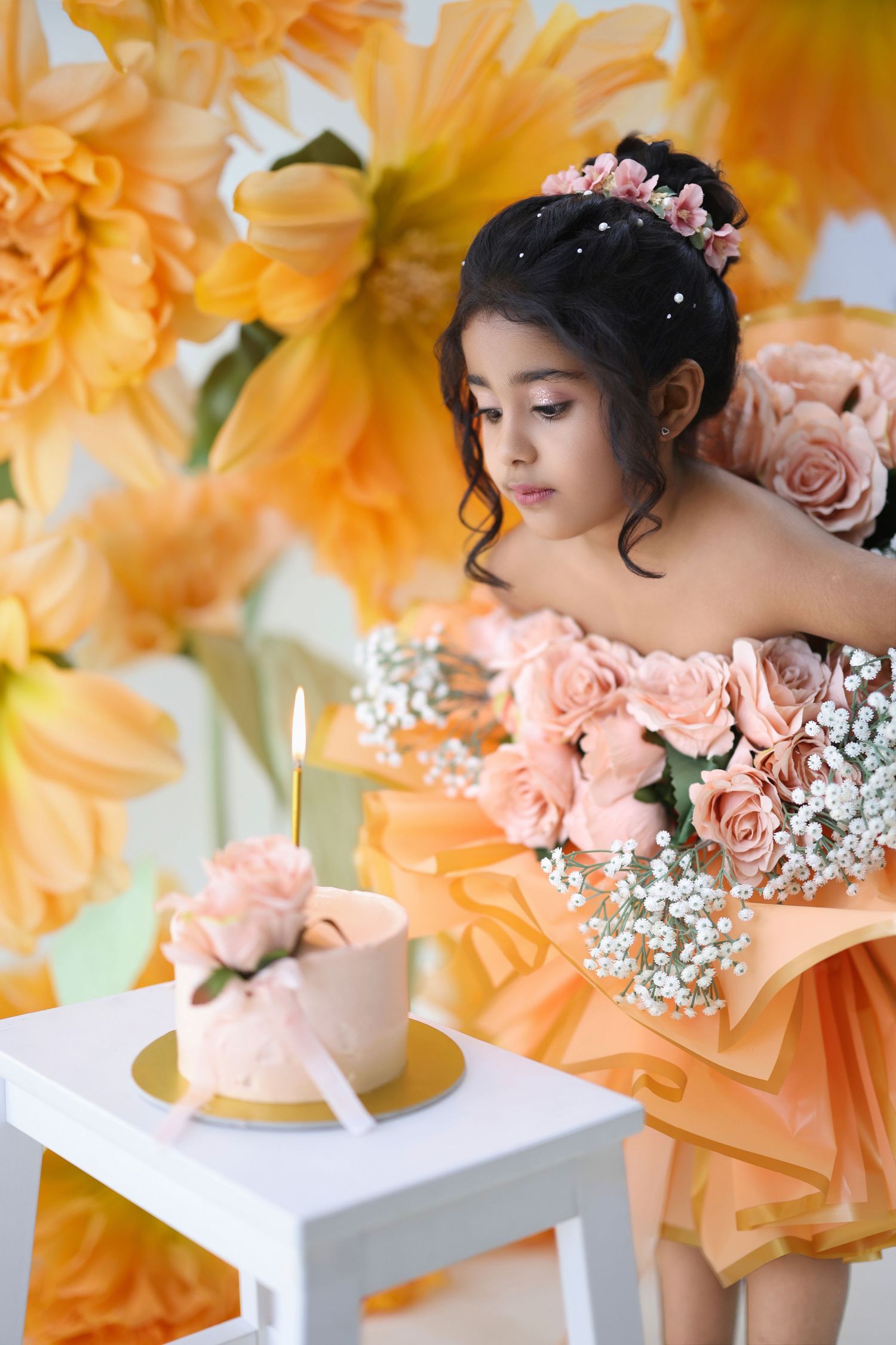 Young girl in peach floral dress with rose bouquet stands before birthday cake with candle amid large yellow flowers.
