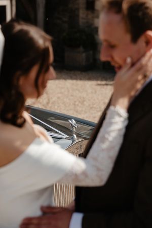 A bride tenderly holds her groom's face near a Rolls-Royce at their wedding venue.