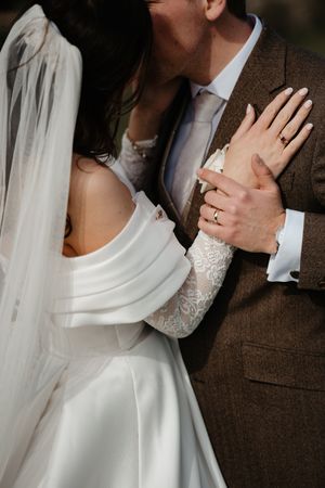 Bride in white off-shoulder gown with lace sleeves embraces groom in brown tweed suit, both wearing wedding rings.