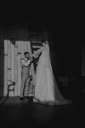Black and white photo of a groom in a vest admiring a wedding dress hanging in a rustic barn.