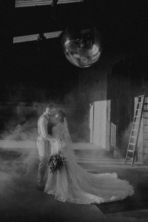 Black and white photo of a bride and groom kissing under a disco ball in a moody, smoke-filled venue.