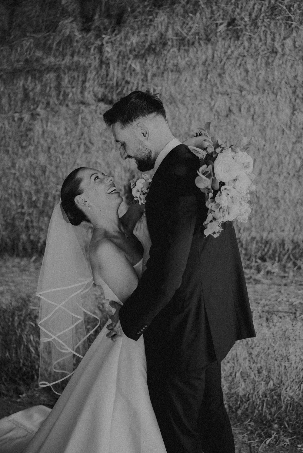 Black and white photo of a laughing bride in a veil and groom in a suit embracing outdoors near hay bales.