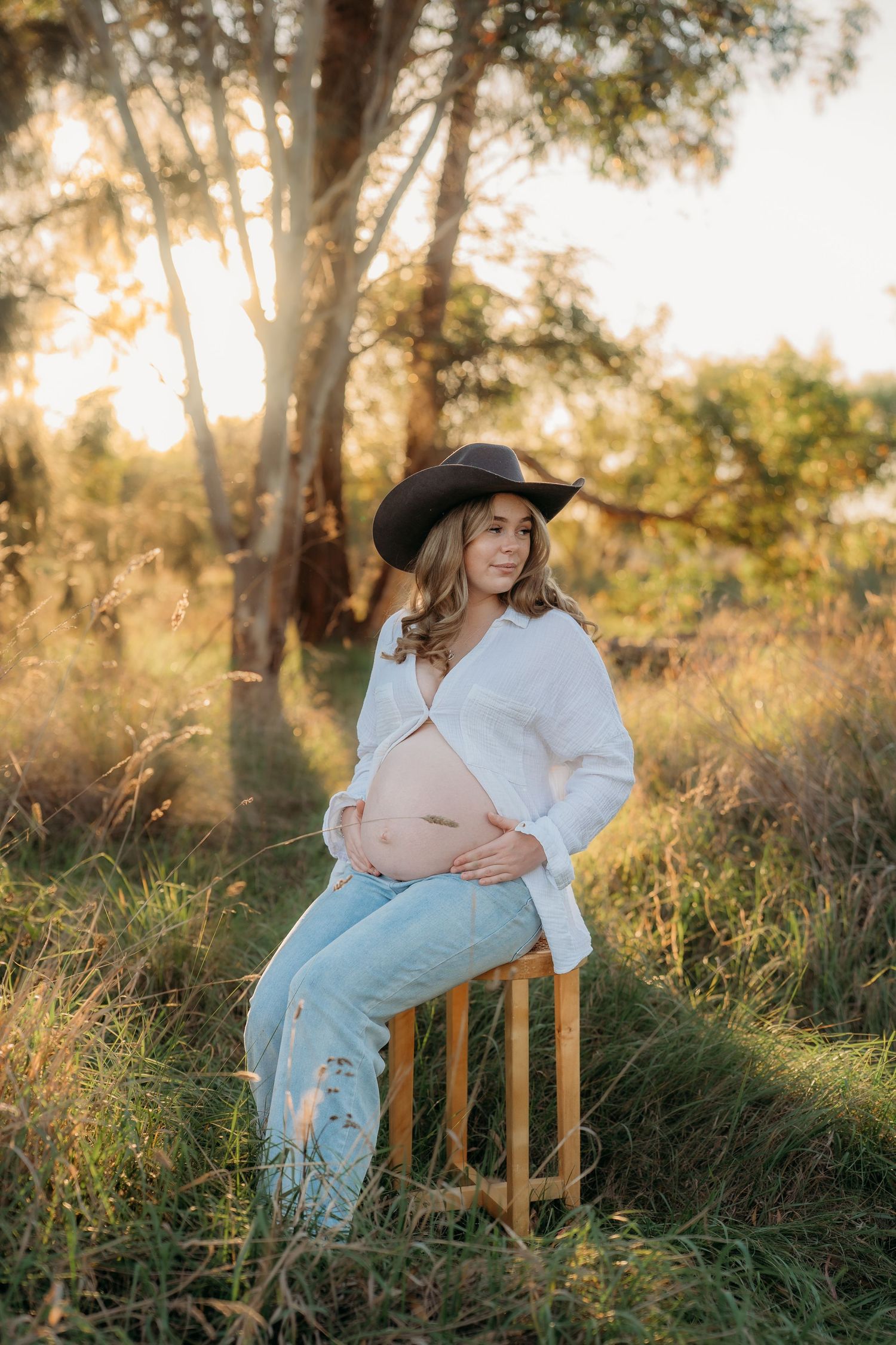 Pregnant woman in cowboy hat and jeans leaning against tree in golden sunset light in a grassy field.