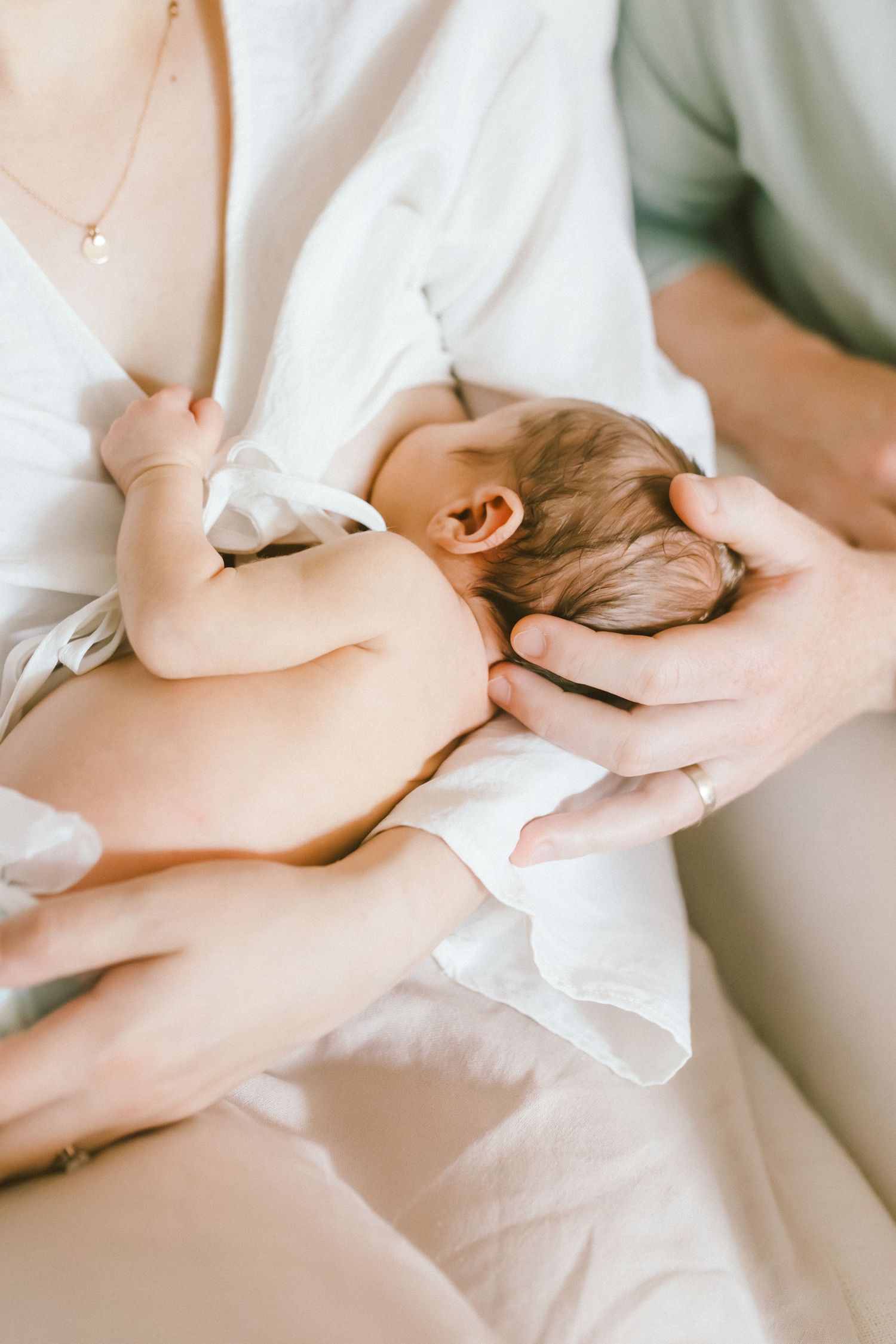 Newborn baby breastfeeding while cradled in mother's arms, with father's hand gently supporting the infant's head.