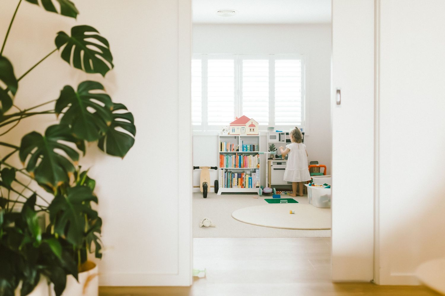 Bright children's playroom viewed through doorway with monstera plant, bookshelf, round rug, and child playing near window.