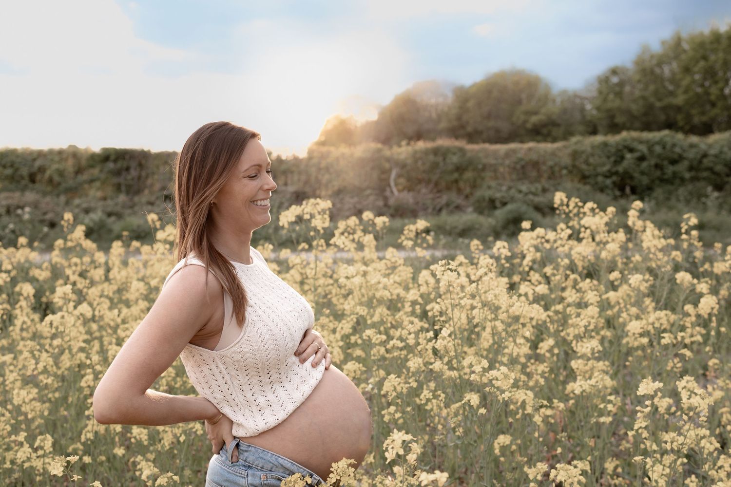 Pregnant woman in white knit top holding baby bump in sunny yellow flower field outdoors.