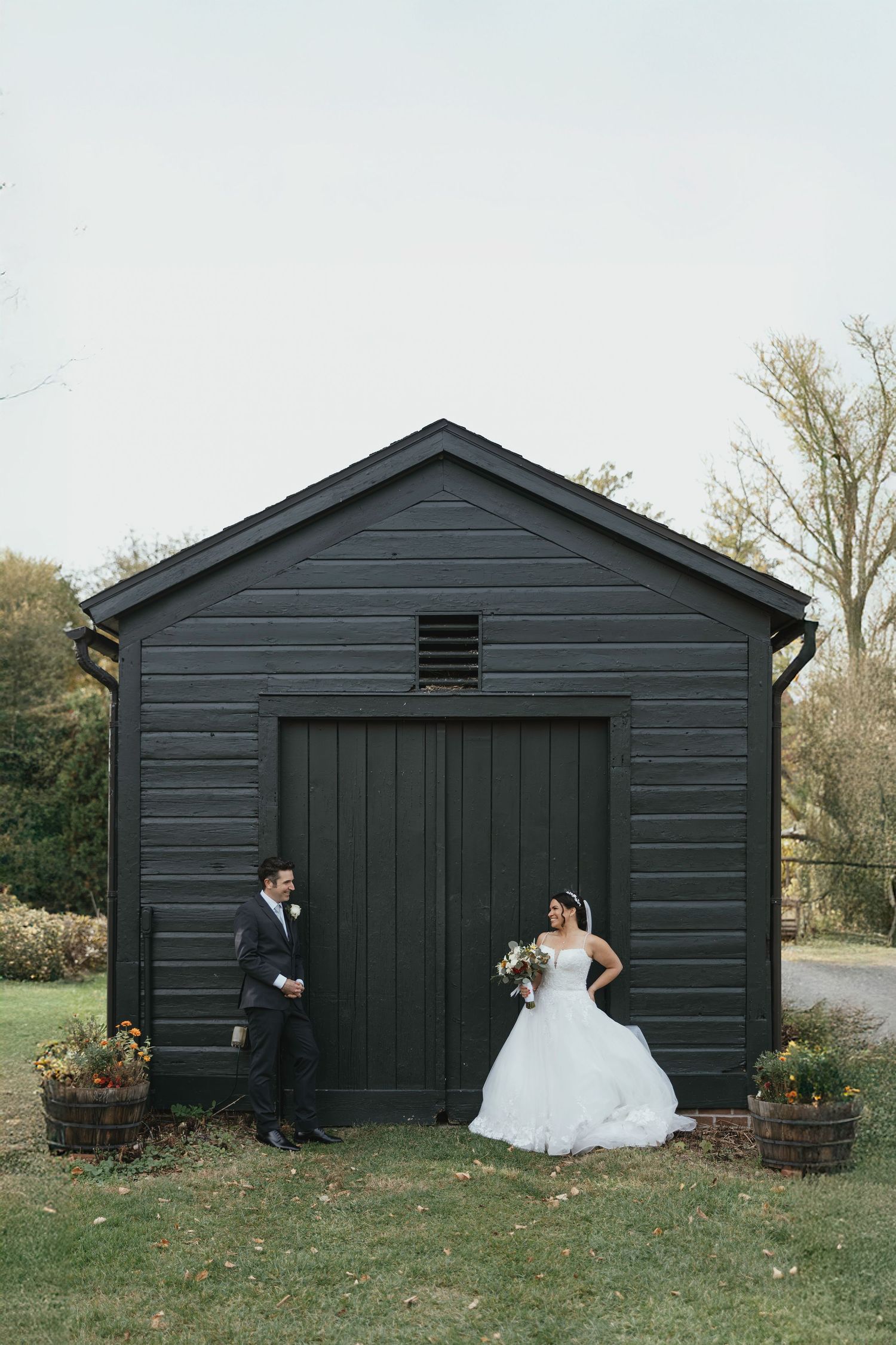 Bride and groom posing in front of a dark green barn on their wedding day in an outdoor autumn setting.