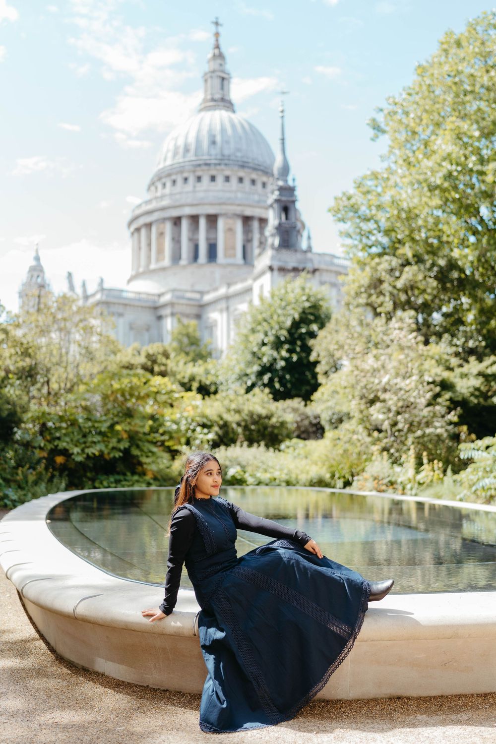 Woman in navy blue dress sitting by a fountain with St. Paul's Cathedral dome visible among trees in London.
