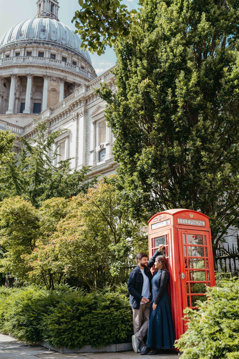 Couple embracing by iconic red phone box near St. Paul's Cathedral, London, surrounded by lush green trees.