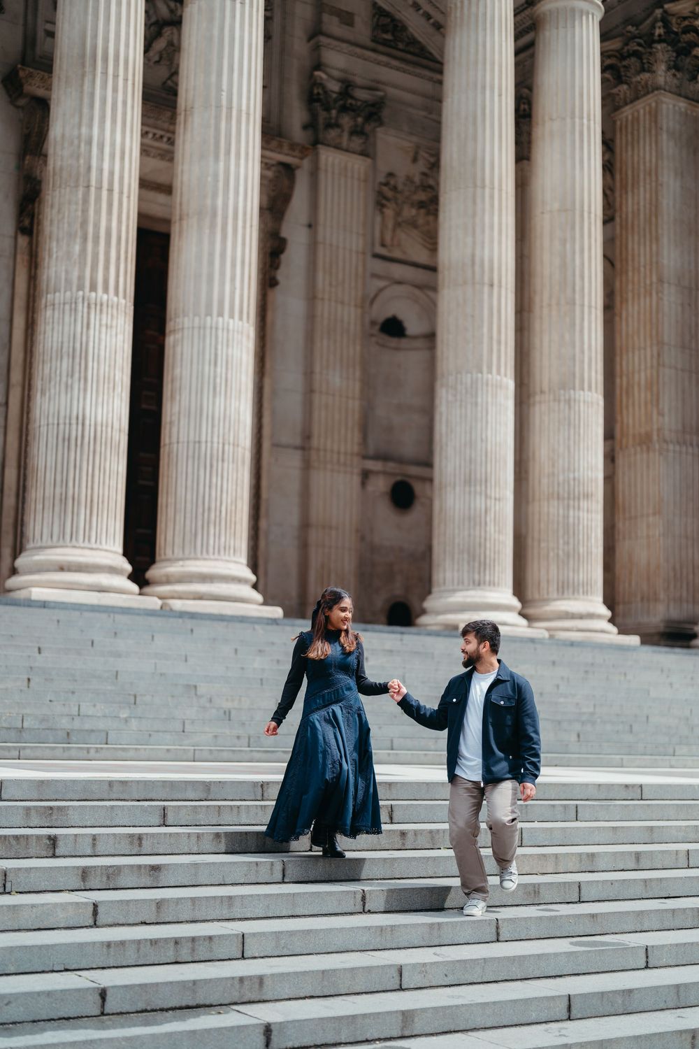 Couple holding hands walking down grand stone steps of a classical building with tall columns.