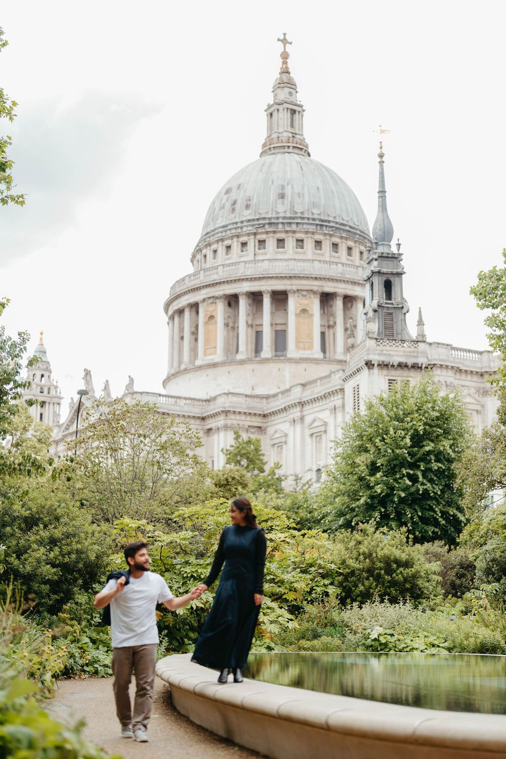 Couple holding hands by a fountain with St. Paul's Cathedral dome and lush greenery in London, England.