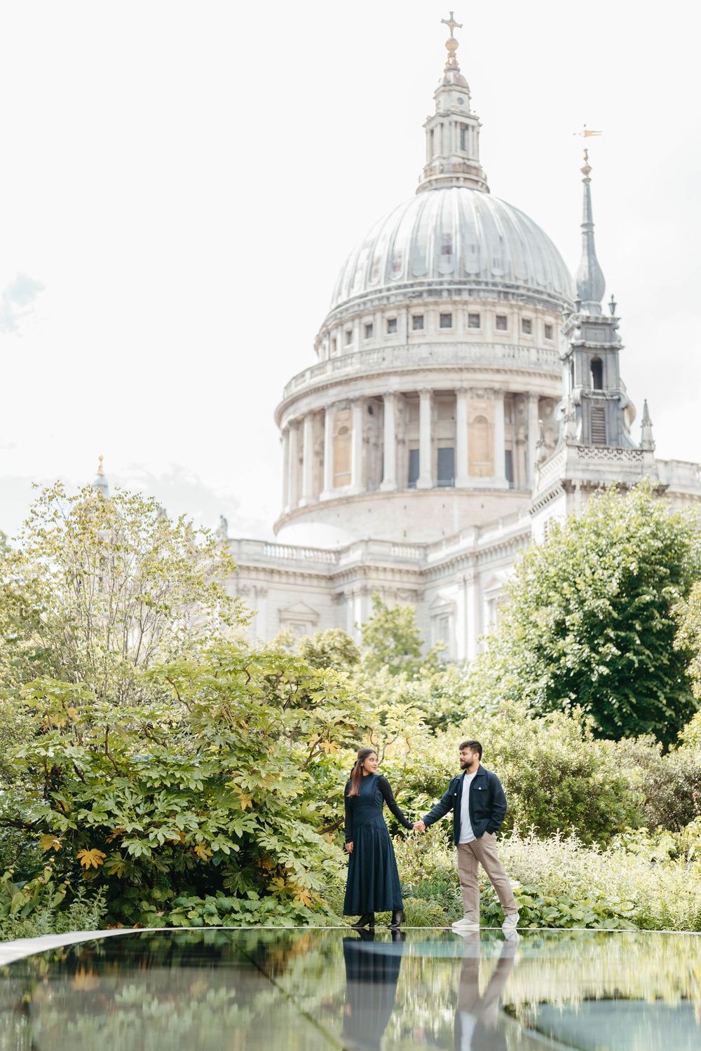 Couple holding hands in front of St. Paul's Cathedral, London, surrounded by lush green trees and gardens.