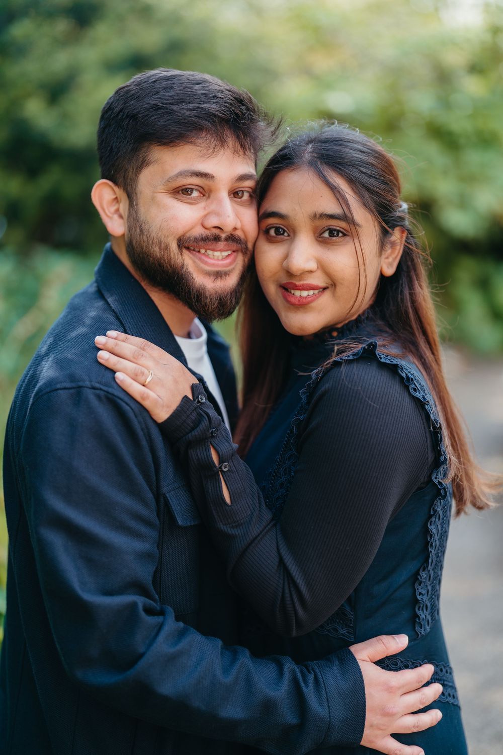 Smiling couple embracing outdoors, man in black jacket hugging woman in dark top, green background.