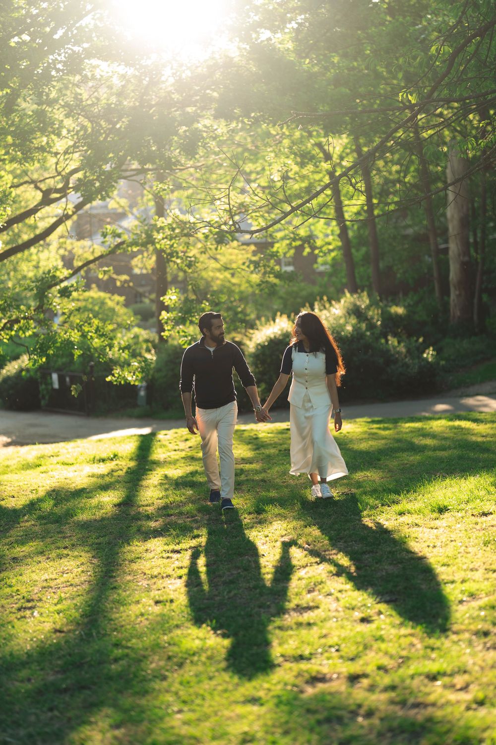 Couple holding hands walking on green grass in sunlit park with long shadows and lush trees in background.
