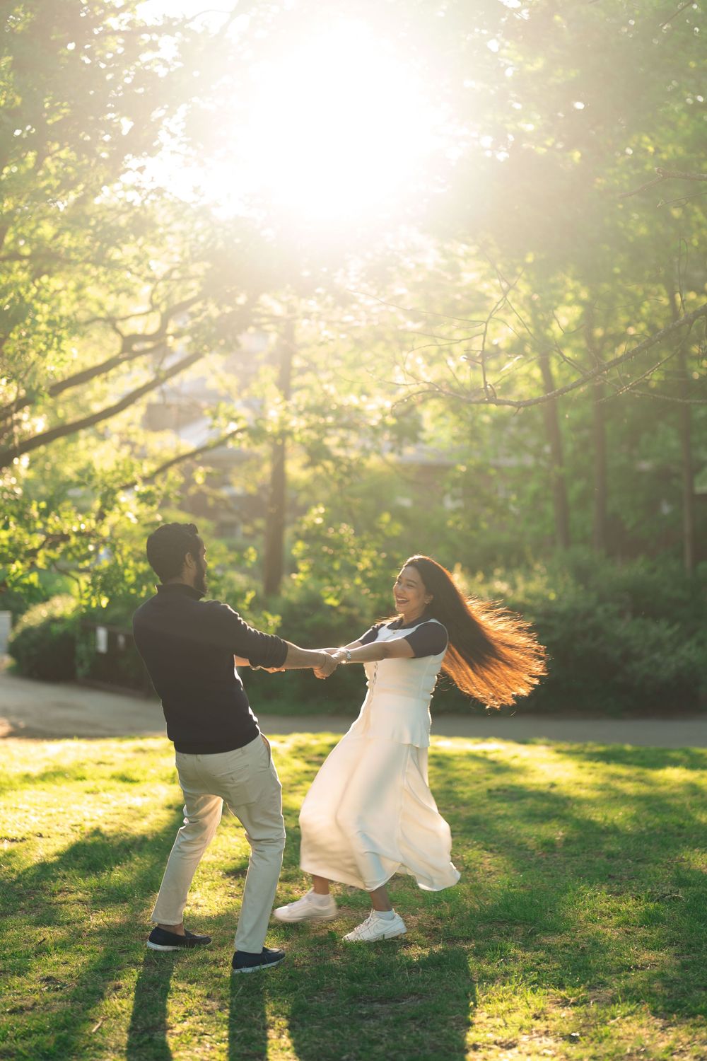 A joyful couple dances and spins together on a sunlit green lawn surrounded by lush trees.