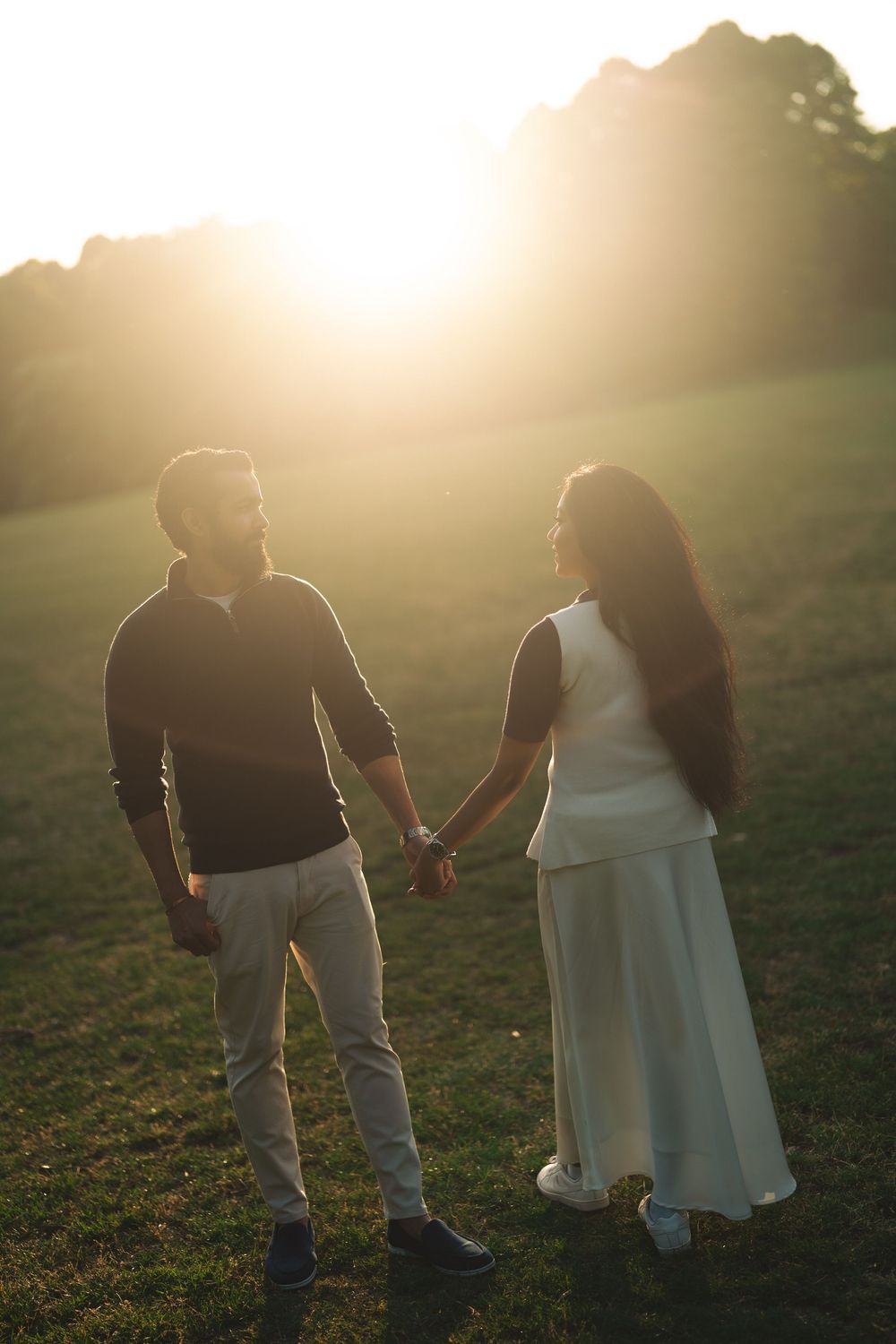 Couple holding hands at golden hour sunset in a green field, man in dark sweater and woman in white dress.