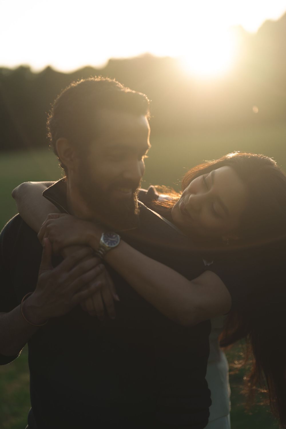 Couple embracing at golden hour sunset in a park, woman smiling with arms around man wearing a watch.