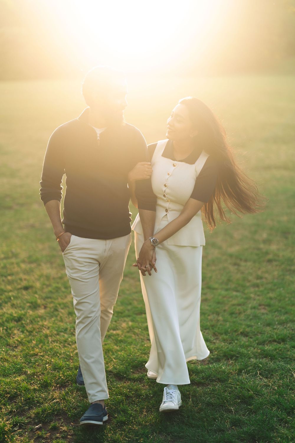 Romantic couple walking hand in hand through a sunlit green field, woman in white dress laughing joyfully.